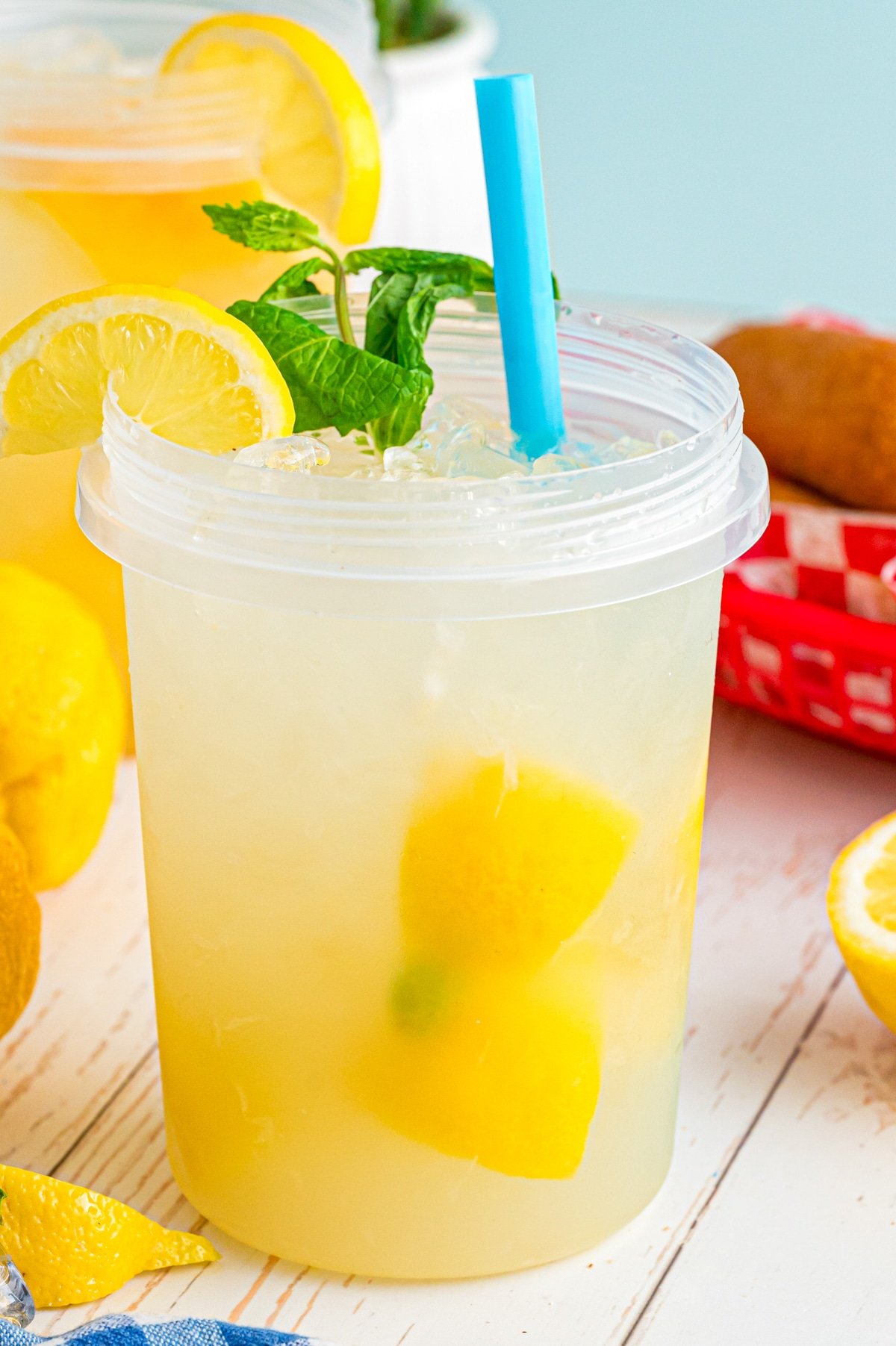 A plastic jar filled with lemonade with a blue straw, a lemon wedge, and mint leaves.