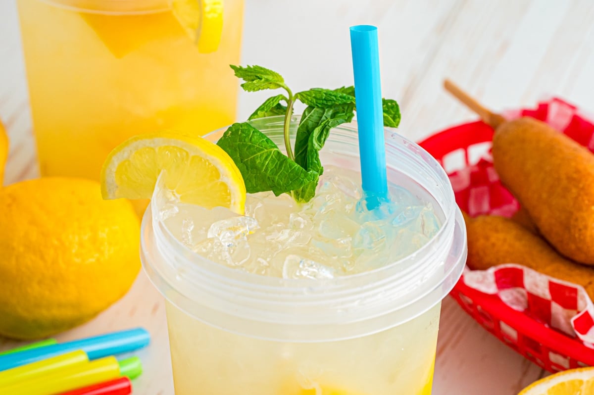 Close-up of the top half of a cup of lemonade showing the top with lemon and mint garnish and a blue straw.